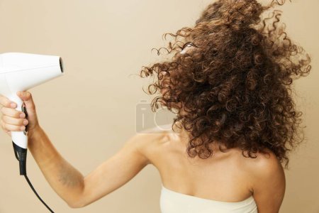 Photo for Woman dries curly hair with blow dryer, home beats styling products, smile on beige background. High quality photo - Royalty Free Image