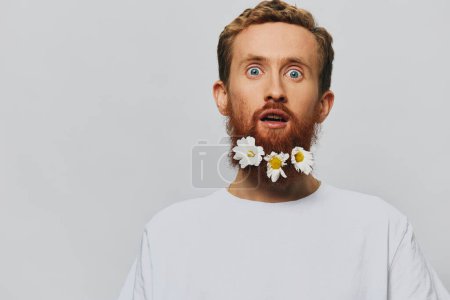 Photo for Portrait of a funny man in a white T-shirt with flowers daisies in his beard on a white isolated background, copy place. Holiday concept and congratulations. High quality photo - Royalty Free Image