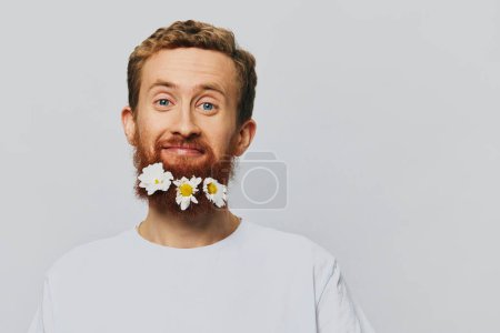 Foto de Retrato de un hombre divertido en una camiseta blanca con flores margaritas en su barba sobre un fondo blanco aislado, lugar de copia. Concepto de vacaciones y felicitaciones. Foto de alta calidad - Imagen libre de derechos