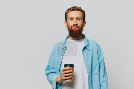 Foto de Hombre hipster con una taza de café con una sonrisa sobre un fondo gris en una camisa azul y camiseta blanca. Foto de alta calidad - Imagen libre de derechos