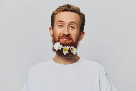Téléchargez les photos : Portrait d'un homme drôle dans un T-shirt blanc avec des marguerites de fleurs dans sa barbe sur un fond blanc isolé, endroit de copie. Concept vacances et félicitations. Photo de haute qualité - en image libre de droit