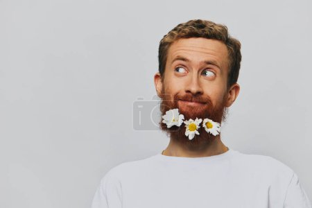 Téléchargez les photos : Portrait d'un homme drôle dans un T-shirt blanc avec des marguerites de fleurs dans sa barbe sur un fond blanc isolé, endroit de copie. Concept vacances et félicitations. Photo de haute qualité - en image libre de droit
