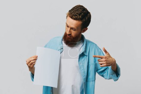 Foto de Retrato de un hombre adulto sobre un fondo gris con una hoja blanca de papel en sus manos para su diseño y texto, diseño, espacio de copia, espacio para texto, señalando con el dedo. Foto de alta calidad - Imagen libre de derechos