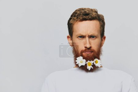Foto de Retrato de un hombre divertido en una camiseta blanca con flores margaritas en su barba sobre un fondo blanco aislado, lugar de copia. Concepto de vacaciones y felicitaciones. Foto de alta calidad - Imagen libre de derechos