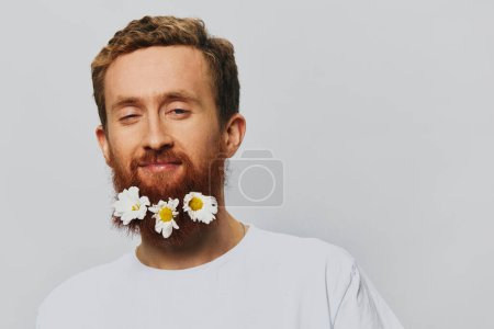 Photo for Portrait of a funny man in a white T-shirt with flowers daisies in his beard on a white isolated background, copy place. Holiday concept and congratulations. High quality photo - Royalty Free Image