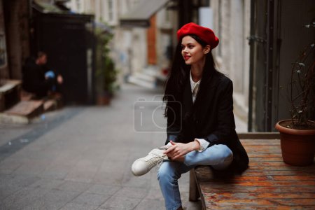 Photo for Stylish woman tourist in stylish clothes in a jacket and red beret sitting outside with phone in hand, cell phone in travel, French style, cinematic color. High quality photo - Royalty Free Image