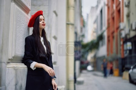 Téléchargez les photos : Femme debout près d'un mur dans la ville portant une veste élégante et béret rouge avec lèvres rouges, Voyage et loisirs, style français de robe, printemps. Photo de haute qualité - en image libre de droit