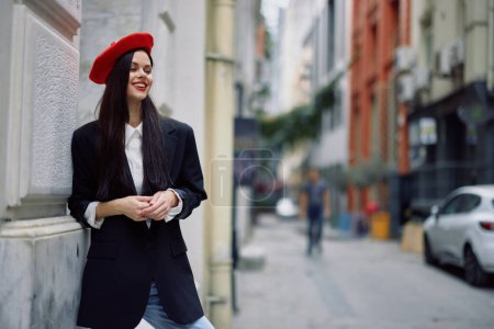 Foto de Mujer de pie cerca de una pared en la ciudad con una chaqueta elegante y boina roja con labios rojos, viajes y ocio, estilo francés de vestido, primavera. Foto de alta calidad - Imagen libre de derechos