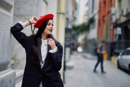 Téléchargez les photos : Une femme debout près d'un mur dans la ville portant une veste élégante et un béret rouge avec des lèvres rouges, voyageant. Photo de haute qualité - en image libre de droit