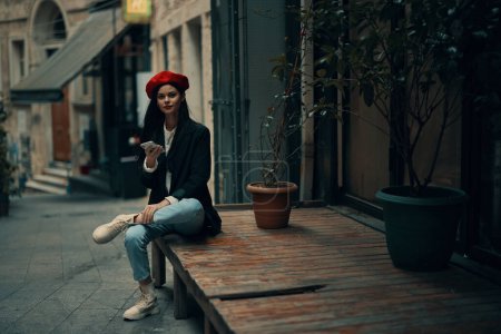 Téléchargez les photos : Femme élégante touriste en vêtements élégants dans une veste et béret rouge assis à l'extérieur avec téléphone à la main, téléphone portable en voyage, style français, couleur cinématographique. Photo de haute qualité - en image libre de droit