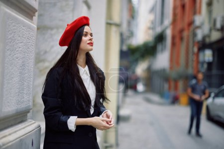 Téléchargez les photos : Une femme debout près d'un mur dans la ville portant une veste élégante et un béret rouge avec des lèvres rouges, voyageant. Photo de haute qualité - en image libre de droit