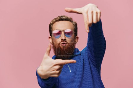 Photo for Portrait of a redheaded man wearing sunglasses smiling and dancing, listening to music on a pink background. Hipster with a beard, happiness finger pointing. High quality photo - Royalty Free Image