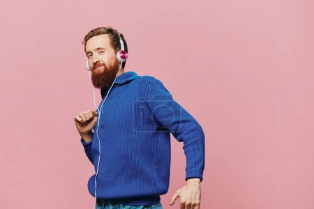 Foto de Retrato de un hombre pelirrojo con auriculares sonriendo y bailando, escuchando música sobre un fondo rosa. Un hipster con barba. Foto de alta calidad - Imagen libre de derechos