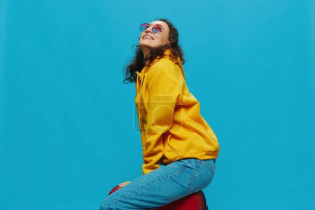 Foto de Mujer sonriente sentada en una maleta en una sudadera con capucha amarilla, jeans azules y gafas sobre un fondo azul, empacando para un viaje. Foto de alta calidad - Imagen libre de derechos