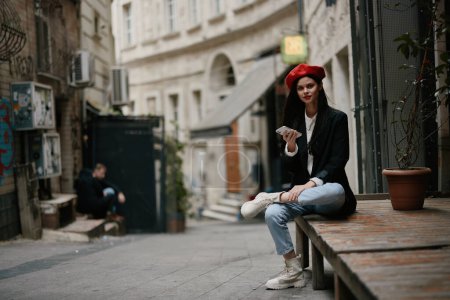 Foto de Mujer turista con estilo en ropa elegante en una chaqueta y boina roja sentado fuera con el teléfono en la mano, teléfono celular en los viajes, estilo francés, color cinematográfico. Foto de alta calidad - Imagen libre de derechos