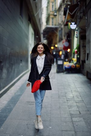Téléchargez les photos : Femme de mode touristique en vêtements élégants dans une veste et béret rouge courant dans la rue étroite de la ville sourire et voyage de bonheur, style français, couleur cinématographique. Photo de haute qualité - en image libre de droit