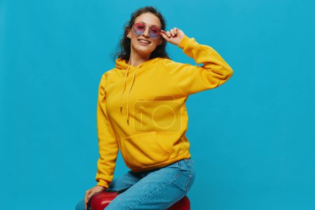 Téléchargez les photos : Femme souriante assise sur une valise avec un sweat à capuche jaune, un jean bleu et des lunettes sur un fond bleu, faisant ses valises pour un voyage. Photo de haute qualité - en image libre de droit