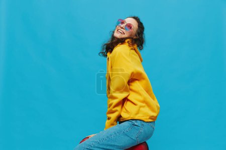 Photo for Smile woman sitting on a suitcase in a yellow hoodie, blue jeans and glasses on a blue background, packing for a trip. High quality photo - Royalty Free Image