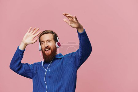 Foto de Retrato de un hombre pelirrojo con auriculares sonriendo y bailando, escuchando música sobre un fondo rosa. Un hipster con barba. Foto de alta calidad - Imagen libre de derechos