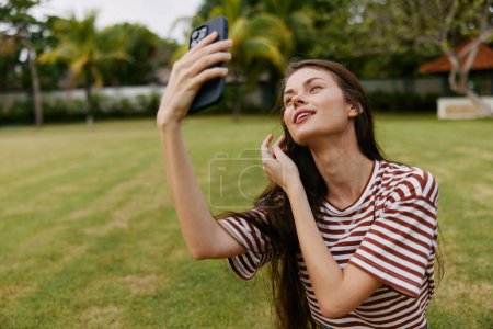 Téléchargez les photos : T-shirt femme nature souriant palmier navigation téléphone extérieur jeans parc marcher vert blogueur style de vie cellulaire herbe d'été à l'extérieur arbre heureux caucasien travail - en image libre de droit