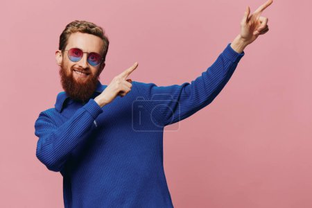 Photo for Portrait of a redheaded man wearing sunglasses smiling and dancing, listening to music on a pink background. Hipster with a beard, happiness finger pointing. High quality photo - Royalty Free Image