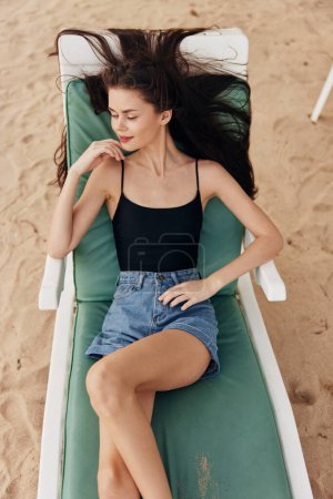 Téléchargez les photos : Femme couché brunette bikini resort chapeau bronzage plage jeune mer assis océan bleu loisirs relax maillot de bain souriant transat femme style de vie sable - en image libre de droit