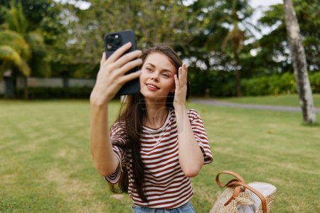 Photo for Woman app palm person blogger grass jeans shirt adult bag smiling tree caucasian nature application call lifestyle smile park happy phone spring - Royalty Free Image