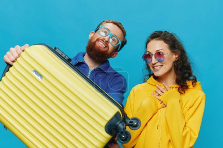 Téléchargez les photos : Femme et homme sourient assis sur une valise avec un sourire jaune, sur fond bleu, emballage pour le voyage, voyage de vacances en famille. Photo de haute qualité - en image libre de droit
