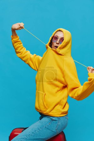 Photo for Smile woman sitting on a suitcase in a yellow hoodie, blue jeans and glasses on a blue background, packing for a trip. High quality photo - Royalty Free Image
