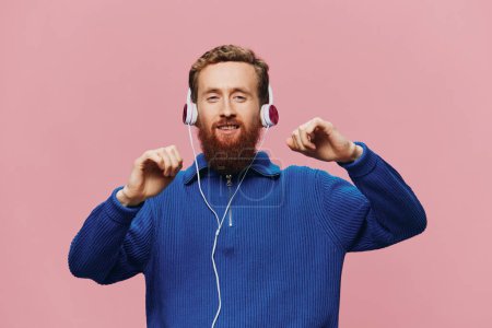Téléchargez les photos : Portrait d'un homme roux portant des écouteurs souriant et dansant, écoutant de la musique sur fond rose. Un hipster avec une barbe. Photo de haute qualité - en image libre de droit