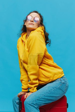 Téléchargez les photos : Femme souriante assise sur une valise avec un sweat à capuche jaune, un jean bleu et des lunettes sur un fond bleu, faisant ses valises pour un voyage. Photo de haute qualité - en image libre de droit