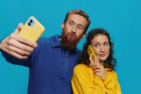 Téléchargez les photos : Femme et homme couple drôle avec des téléphones à la main prenant selfies sourire tordu amusant, sur fond bleu. Le concept de vraies relations familiales, parler au téléphone, travailler en ligne. Photo de haute qualité - en image libre de droit