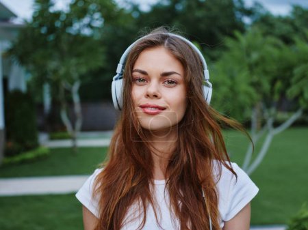 Téléchargez les photos : Femme portant un casque écoutant de la musique sourire avec des dents regardant vers la caméra en t-shirt blanc, voyageant en Asie. Photo de haute qualité - en image libre de droit