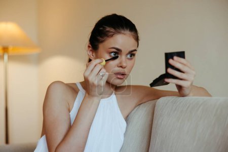 Téléchargez les photos : Une femme à la maison regarde dans le miroir et applique des cils de mascara de colorant de maquillage, concept de beauté, soin de la peau à problème avec l'acné. Photo de haute qualité - en image libre de droit