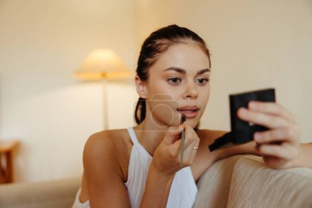 Téléchargez les photos : Une femme à la maison se regarde dans le miroir et applique du rouge à lèvres de maquillage, concept de beauté, soin de la peau problème avec l'acné, maquillage à la maison. Photo de haute qualité - en image libre de droit