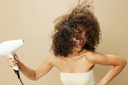 Foto de Mujer seca el cabello rizado con secador de pelo, casa late productos de peinado, sonrisa sobre fondo beige. Foto de alta calidad - Imagen libre de derechos