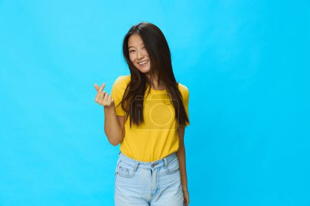 Photo for Asian woman smiling with teeth pointing finger at loose background in yellow T-shirt and blue jeans on blue background, copy space. High quality photo - Royalty Free Image