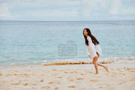 Téléchargez les photos : Femme sportive court le long de la plage en vêtements d'été sur le sable dans un T-shirt jaune et short en jean chemise blanche volant cheveux vue sur l'océan, vacances à la plage. Photo de haute qualité - en image libre de droit