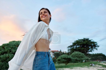 Foto de Mujer morena con el pelo largo en una camisa blanca y pantalones cortos de jean bronceado abdominales y felicidad sonrisa divertida con dientes en la playa y palmeras, vacaciones viaje de verano puesta de sol cielo. Foto de alta calidad - Imagen libre de derechos