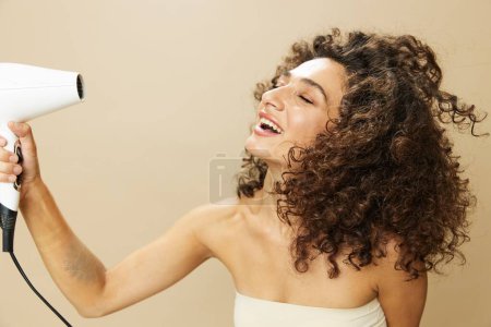 Téléchargez les photos : Femme sèche les cheveux bouclés avec sèche-cheveux, maison bat des produits coiffants, sourire sur fond beige. Photo de haute qualité - en image libre de droit
