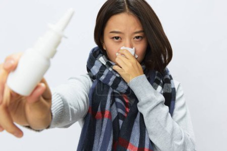 Téléchargez les photos : Femme asiatique avec un rhume avec un nez bouché tenant un spray nasal pour les allergies avec un mal de gorge portant une écharpe à carreaux sur un fond blanc. Photo de haute qualité - en image libre de droit