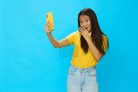 Téléchargez les photos : Heureuse femme asiatique tenant son téléphone et regardant l'écran prendre des selfies avec un boîtier jaune sur un fond bleu dans un T-shirt jaune souriant avec des dents. Photo de haute qualité - en image libre de droit