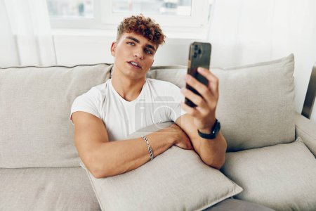 Téléchargez les photos : Homme mâle selfies maison internet jeune sourire tenant cyberespace étudiant sport téléphone portable personne canapé pigiste - en image libre de droit