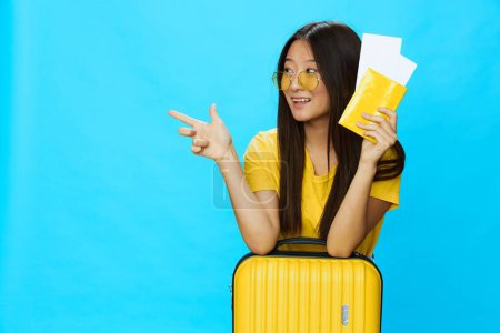 Téléchargez les photos : Femme asiatique voyageant avec une valise jaune et des billets avec passeport à la main, touriste voyageant en avion et en train avec des bagages sur fond bleu. Photo de haute qualité - en image libre de droit