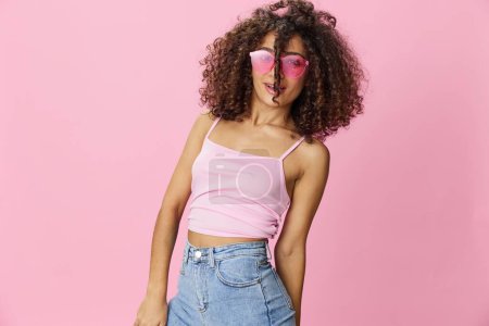 Téléchargez les photos : Happy woman afro boucles cheveux danse sur un fond rose en t-shirt rose d'été jeans et lunettes, ambiance d'été, espace de copie. Photo de haute qualité - en image libre de droit