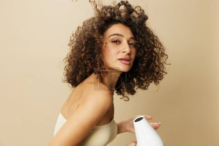 Téléchargez les photos : Femme sèche les cheveux bouclés avec sèche-cheveux, maison bat des produits coiffants, sourire sur fond beige. Photo de haute qualité - en image libre de droit