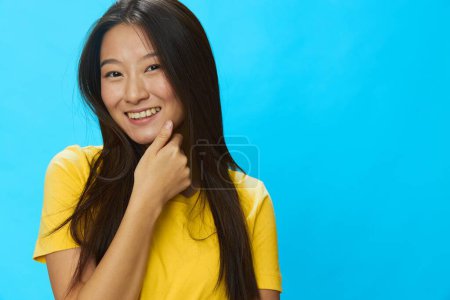 Photo for Asian woman smiling with teeth pointing finger at loose background in yellow T-shirt and blue jeans on blue background, copy space. High quality photo - Royalty Free Image