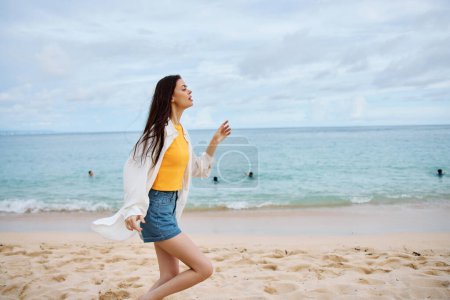Téléchargez les photos : Femme sportive court le long de la plage en vêtements d'été sur le sable dans un débardeur jaune et short en jean chemise blanche volant cheveux vue sur l'océan, vacances à la plage et liberté de voyage. Photo de haute qualité - en image libre de droit