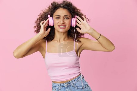 Téléchargez les photos : Femme heureuse portant des écouteurs avec des cheveux bouclés écoutant de la musique et dansant dans un T-shirt rose et un jean sur un fond rose, copiez l'espace. Photo de haute qualité - en image libre de droit