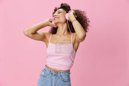 Foto de Mujer feliz usando auriculares con el pelo rizado escuchando música y bailando en una camiseta rosa y jeans sobre un fondo rosa, espacio para copiar. Foto de alta calidad - Imagen libre de derechos